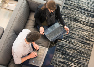 A man and woman sit on a couch with laptops discussing work.